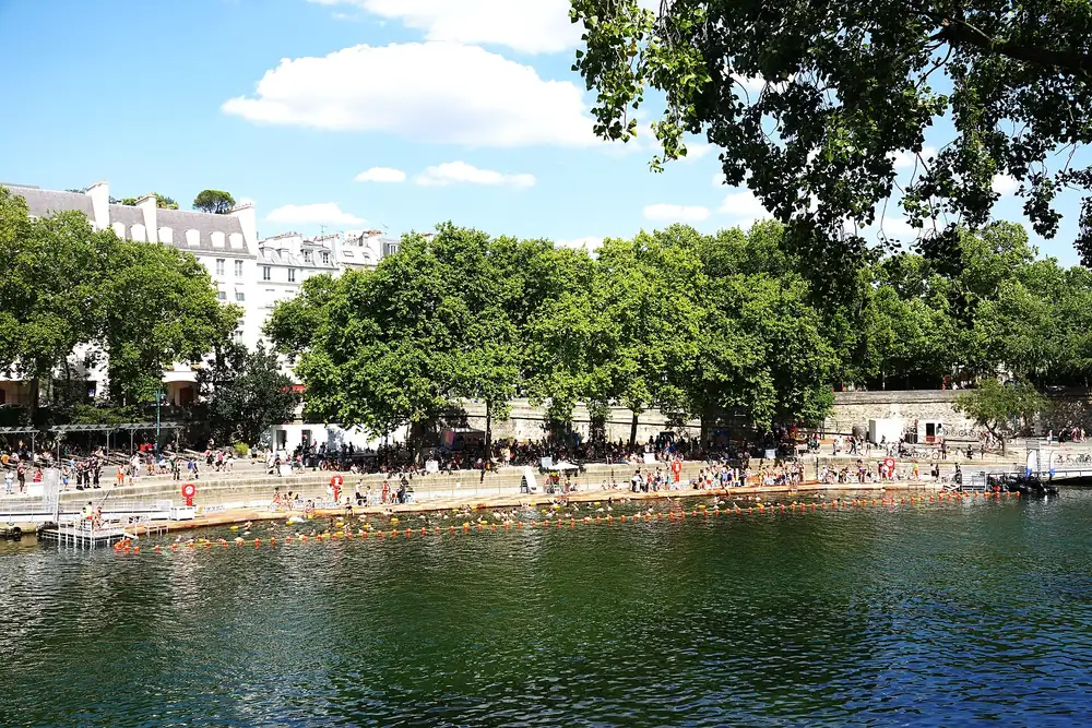 Le bassin de baignade du bras Marie, délimité par des bouées orange le long du quai, avec les baigneurs sur le ponton et les arbres du quai de l'Hôtel-de-Ville