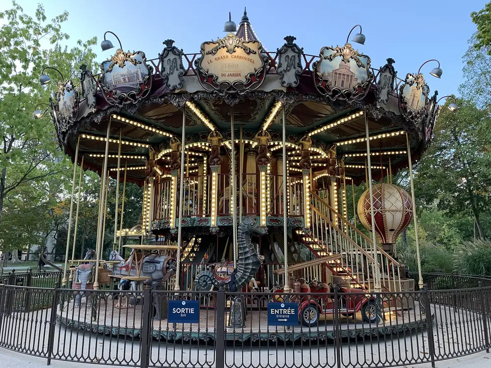 Un carrousel à deux étages du Jardin d'Acclimatation, avec ses lampes, ses chevaux et une montgolfière décorative, derrière une grille noire marquée entrée et sortie