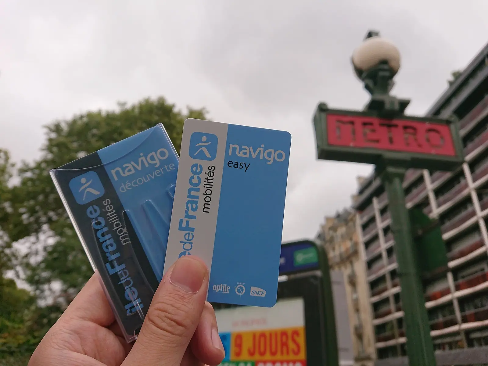 Un Navigo Découverte et un Navigo Easy tenus à la main devant une enseigne Métro